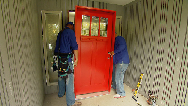 red entry door being installed by men in blue long-sleeve shirts on grey porch