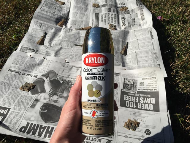 hand holding can of gold spray paint over drawer hardware pulls laying on newspaper in grass